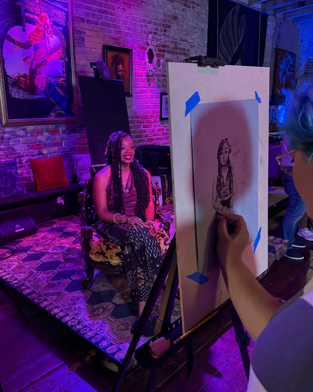 A live artist sketches a portrait of a woman posing in a purple-lit studio.