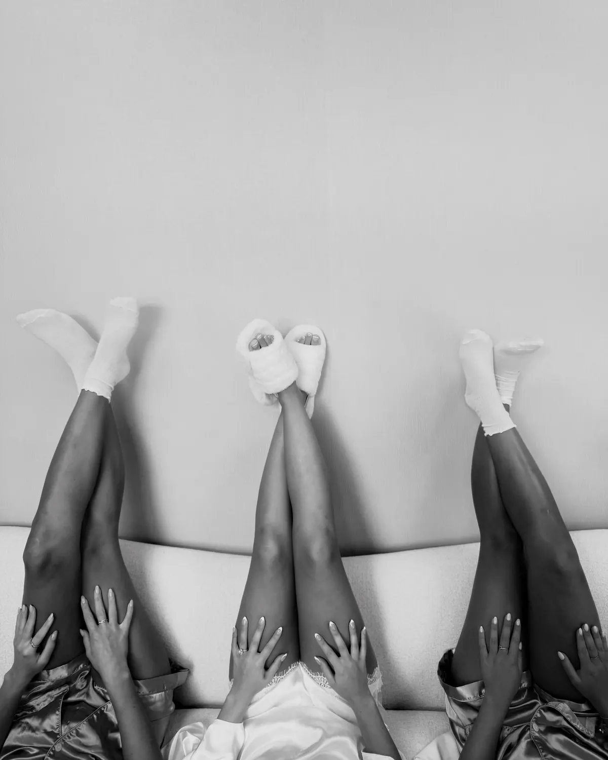 Black-and-white photo of three women lying on a sofa with their legs up the wall in white socks.