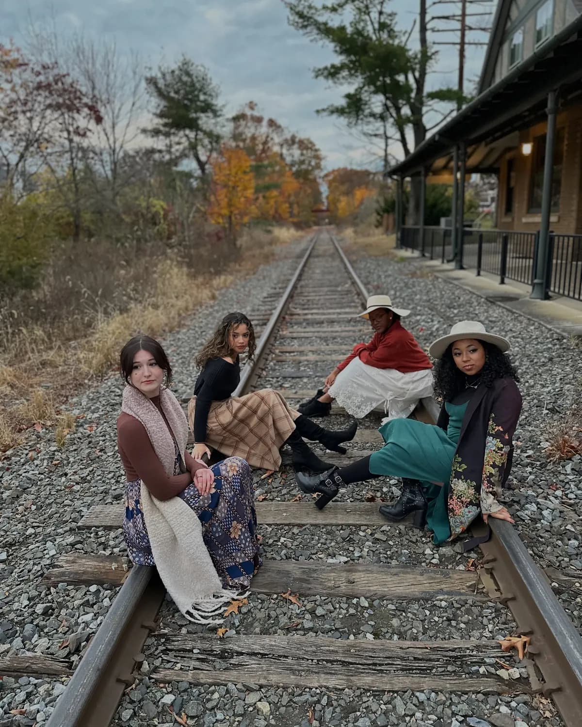 A group of women in vintage-inspired autumn outfits sit together on railroad tracks.